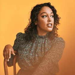 Headshot of Scholar of Note Dr. Eve L. Ewing. She is looking to the side while sitting in a wooden chair in front of a yellow background. Her curly hair is down and she is wearing a long sleeve floral patterned top.