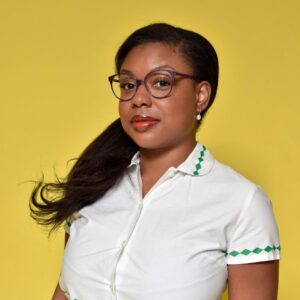 Headshot of the Literacy is Our Legacy Writers Award Winner Kendra Valentine. She is looking at the camera in front of a yellow background, wearing a white button up shirt and glasses. Her long hair is tied to the side.