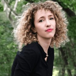 Headshot of Scholar of Note Lilly Dancyger. She is looking at the camera in front of a blurred background of greenery. Her blonde curly hair is down and she is wearing a black top.