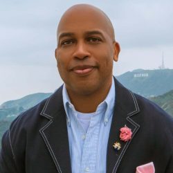 Headshot of Visiting Fellow Rasheed Newson. He is looking at the camera in a blue button down and navy blue jacket with a pink flower pin and gold bug pin on it. You can see the "Hollywood" sign in Los Angeles in the background. The sky is gray.